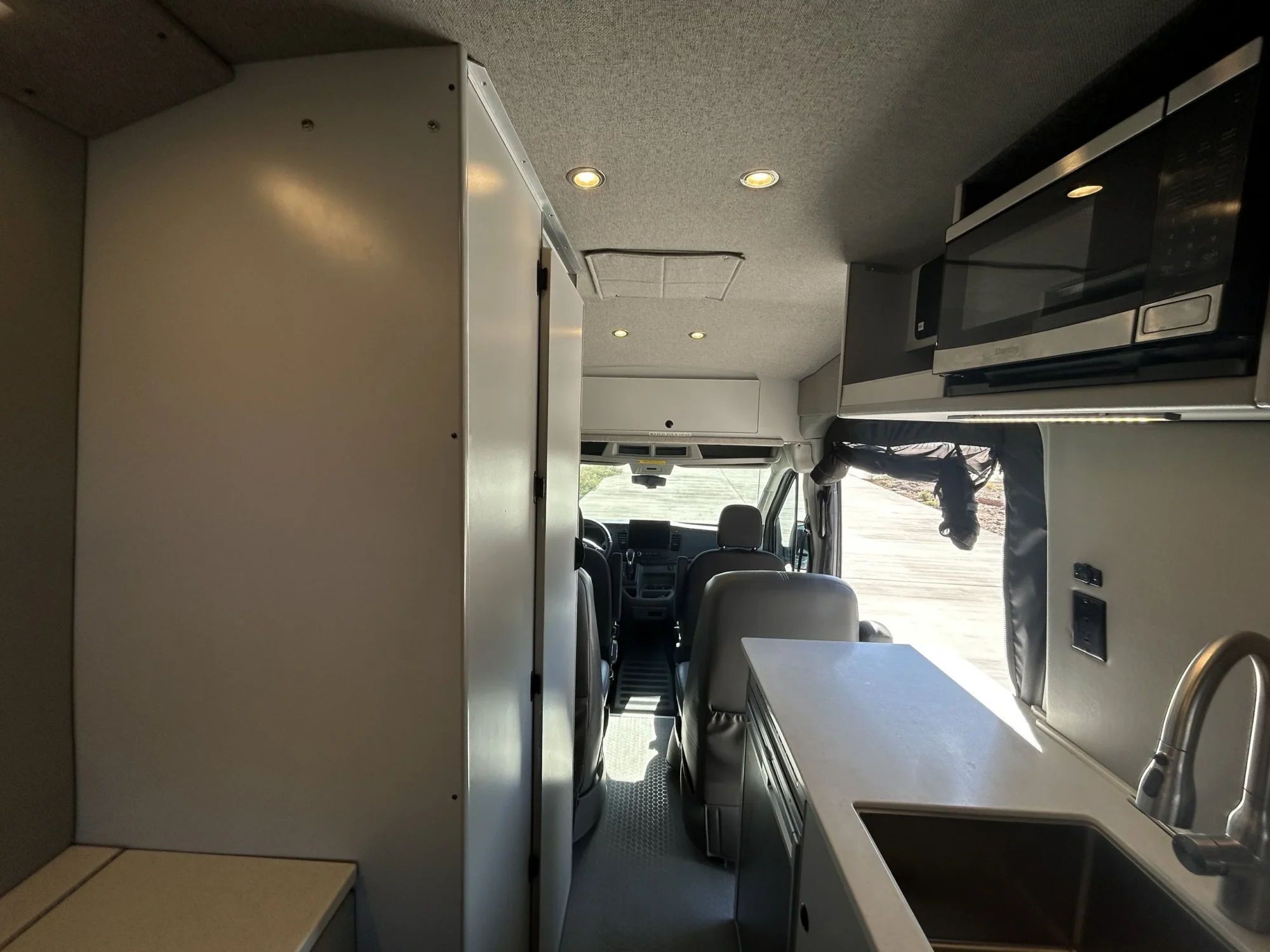 2024 Ford Transit 350 148 High Roof AWD, view from rear toward cab showing galley sink, microwave, bath enclosure, and captain's chairs
