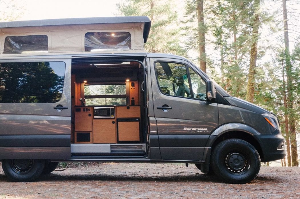 Grey Sportsmobile camper van with a penthouse top with the door open in the woods.