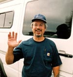 Sportsmobile owner, Gilbert Parklett, stands next to his custom white conversion van waving to the camera.