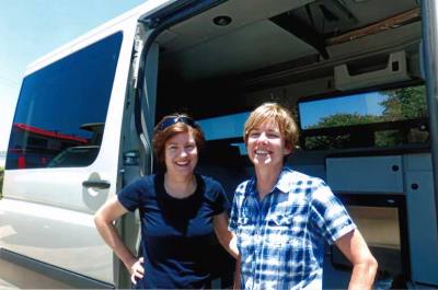 Lady Sportsmobile owner, Sannie Mueller, stands with her friend next to her custom white Sportsmobile conversion van.