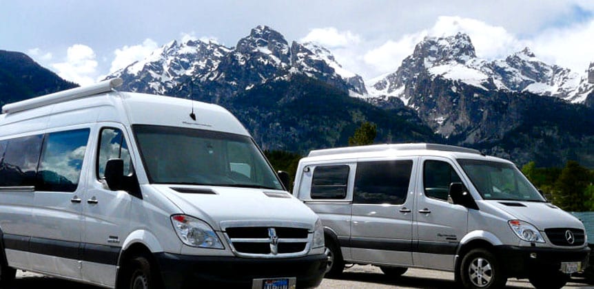 A pair of Sportsmobile custom camper vans with snowy peaks in the background.