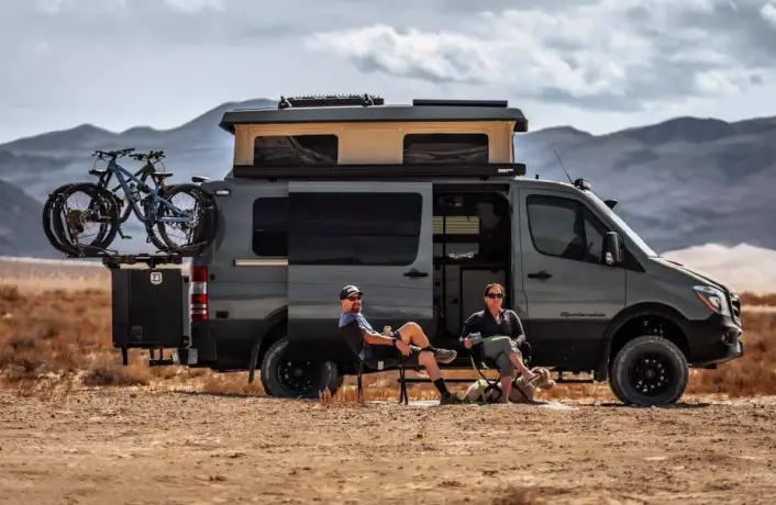 Dark grey Mercedes Sprinter with popped up roof and bikes attached to it's back and two people sitting in the chairs in front of it.