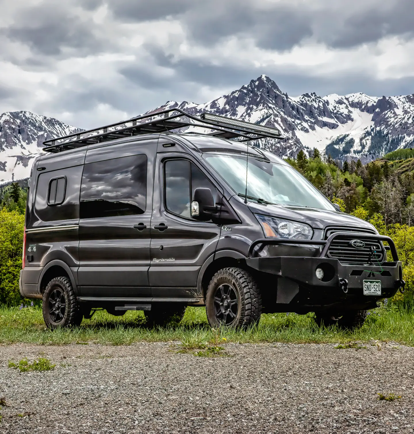 Black van parked in the field. Forest and mountains in the background.