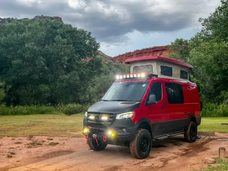 Red Mercedes Sprinter 4x4 camper van with the pop-top raised and exterior lights on
