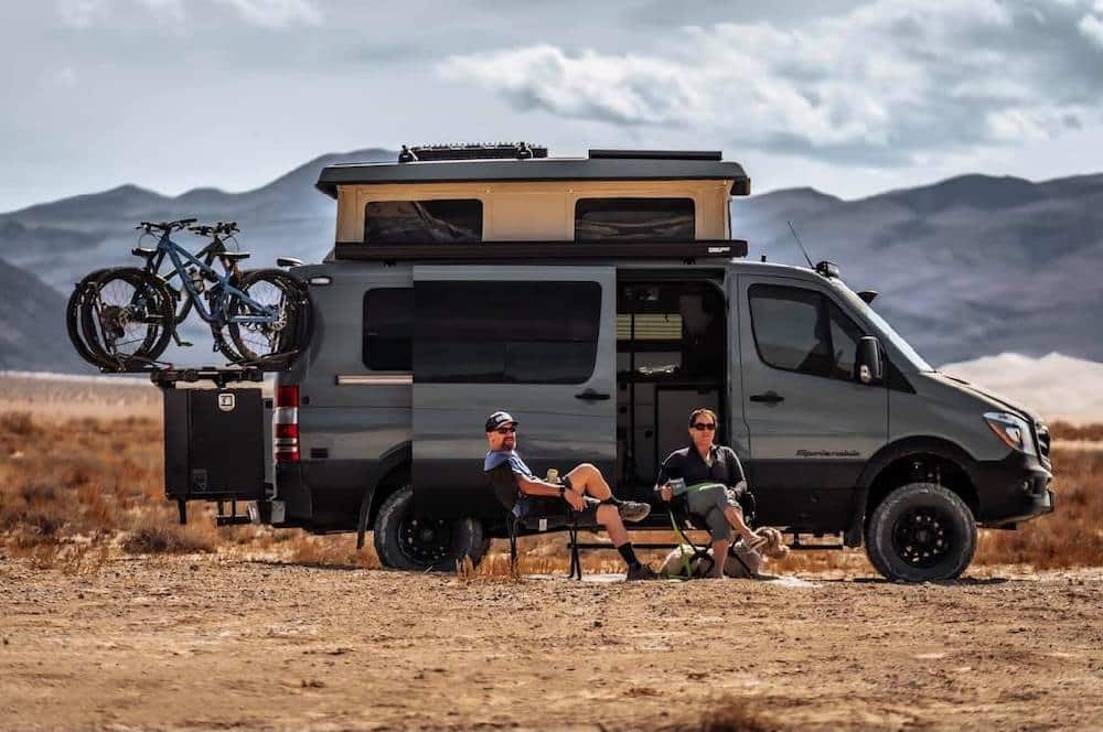 Dark grey Converted Mercedes Sprinter with bikes attached to it's back and two people sitting in the chairs in front of it.