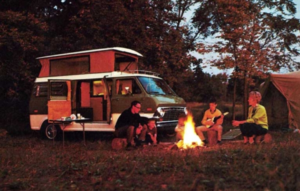 A family of four sitting around the campfire. Their van parked in the forest.
