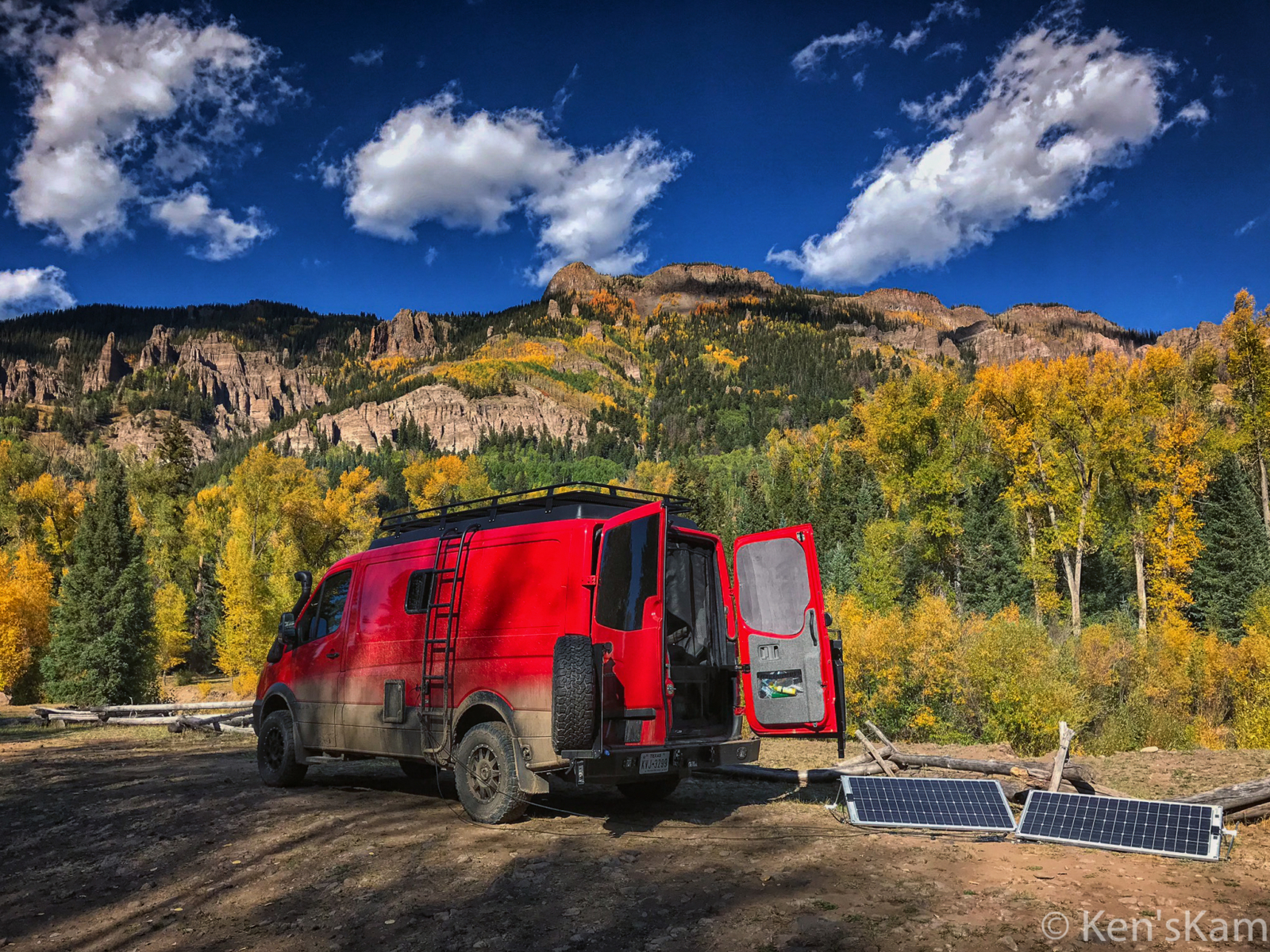 Red converted van with opened rear screen parked next to solar panels. Trees and mountains in the background.