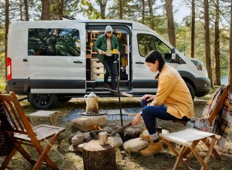 Van parked in the forest. Person tending a campfire while the other person brings food from the van.
